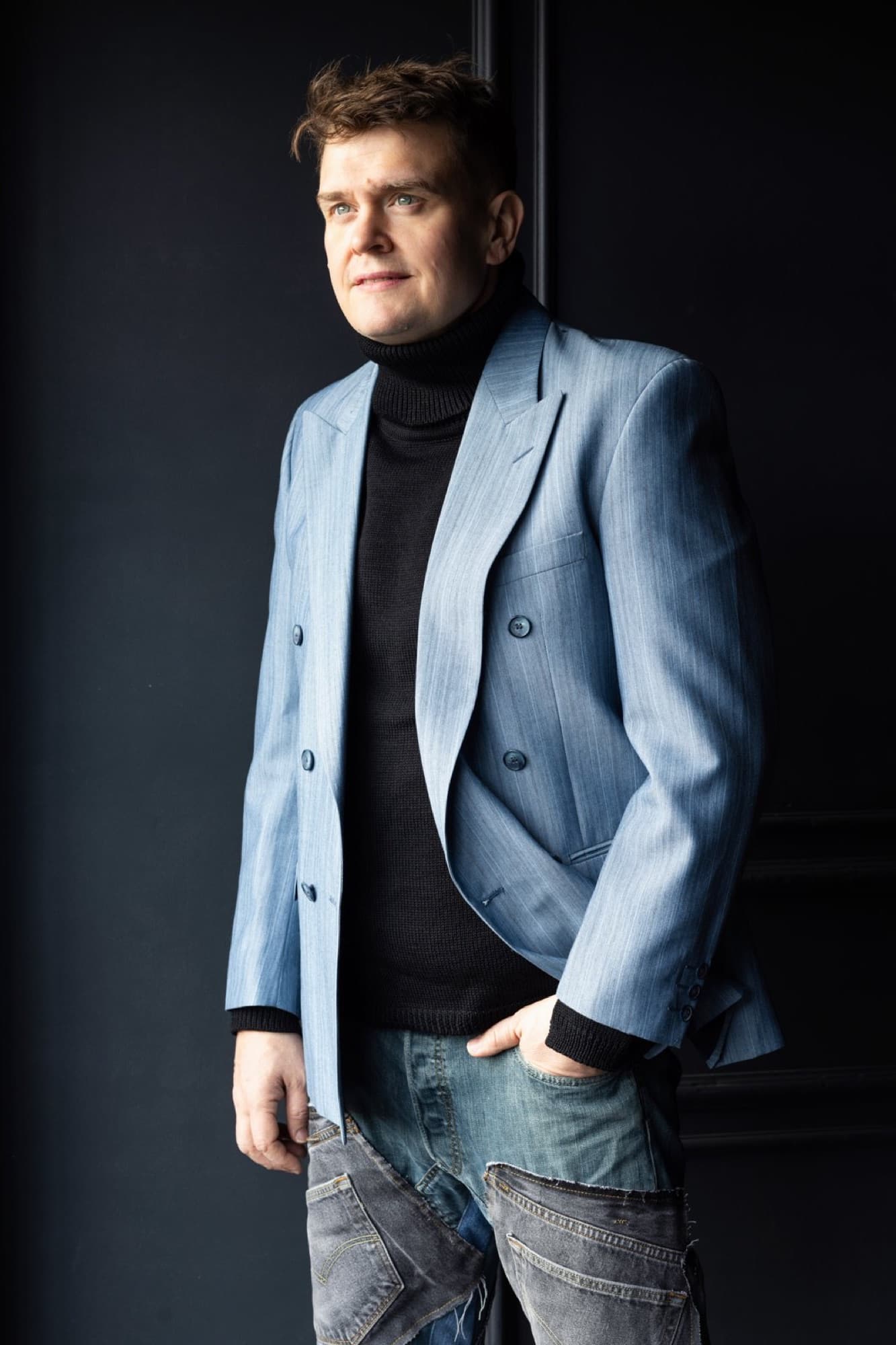 A man stands leaning against a dark wall, wearing a pale blue blazer over a black polo shirt, one hand in his jeans pocket, looking to the side against a dark background.